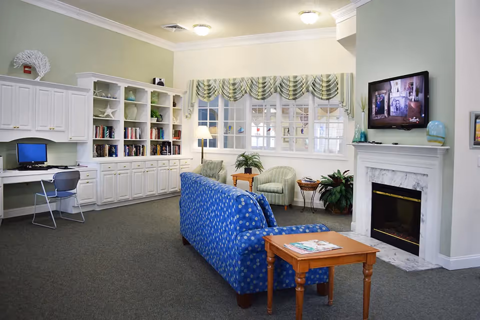 A cozy living room area in a senior living facility featuring a blue patterned couch, a wooden side table with magazines, two green striped armchairs, a white fireplace with a mounted flat-screen TV above it, built-in white cabinetry with books and decorative items, a desk with a computer, and a window with green and white valance curtains.