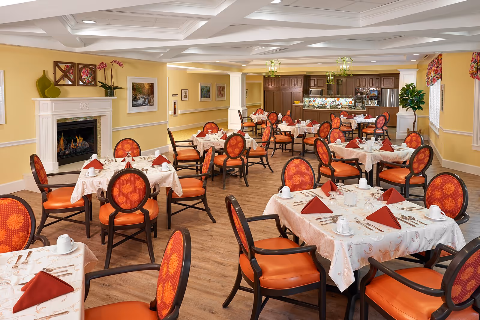 A bright and elegant dining room with multiple tables covered in white tablecloths and set with red folded napkins, white cups, and silverware. The chairs have orange cushions and patterned backs. The room features yellow walls, a fireplace with decorative vases and flowers on the mantel, framed artwork, and a kitchen area with dark wood cabinets and colorful backsplash in the background.