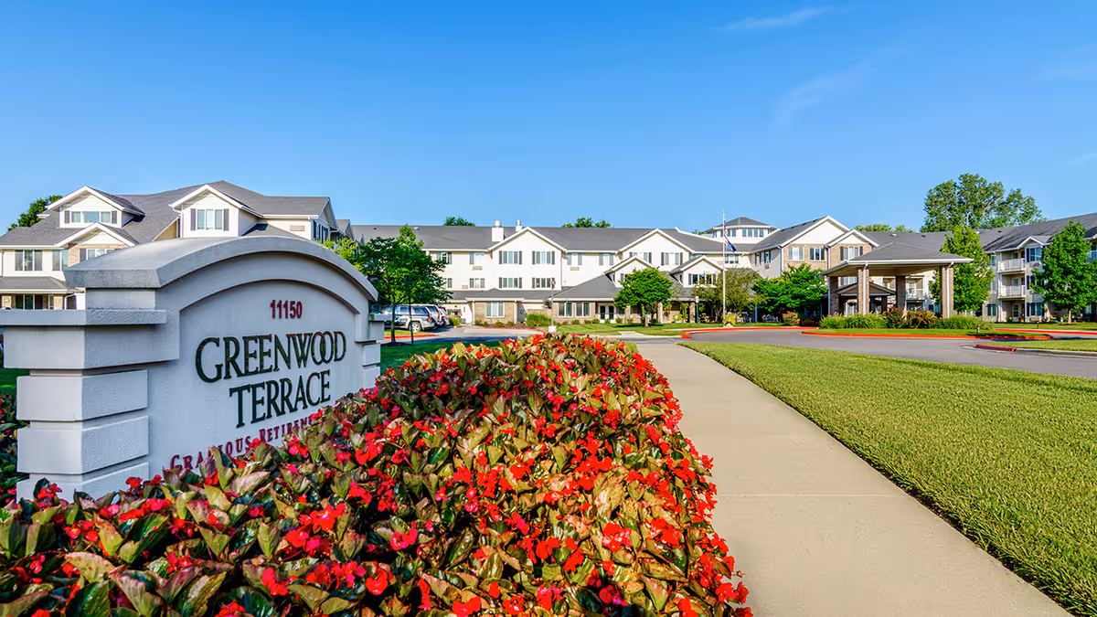 Exterior view of Greenwood Terrace senior living facility with a large sign in the foreground surrounded by red flowers, a paved walkway, green lawn, and a multi-story building under a clear blue sky.