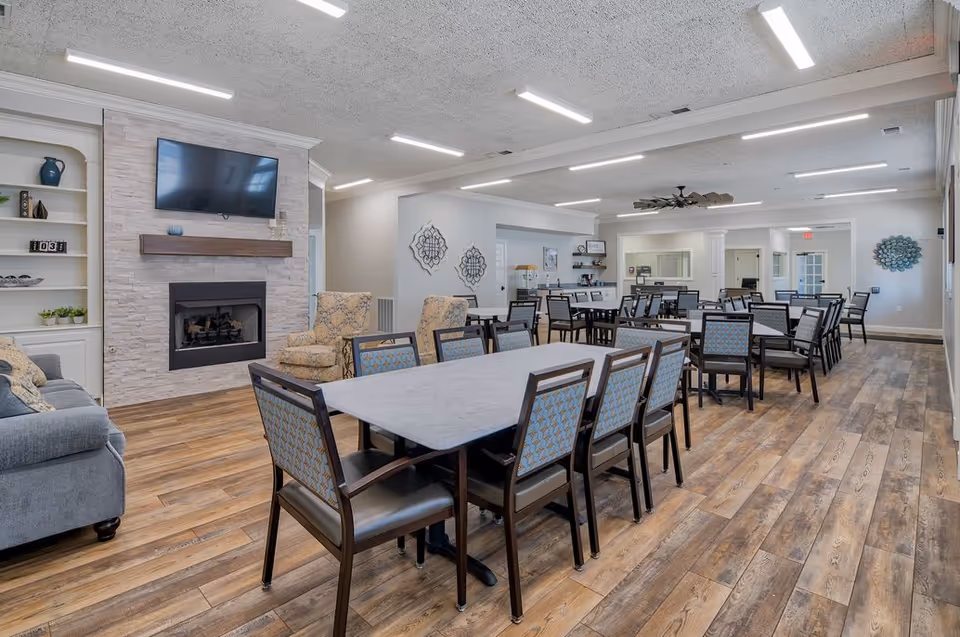 Bright senior living dining hall with long tables, chairs, and a fireplace.