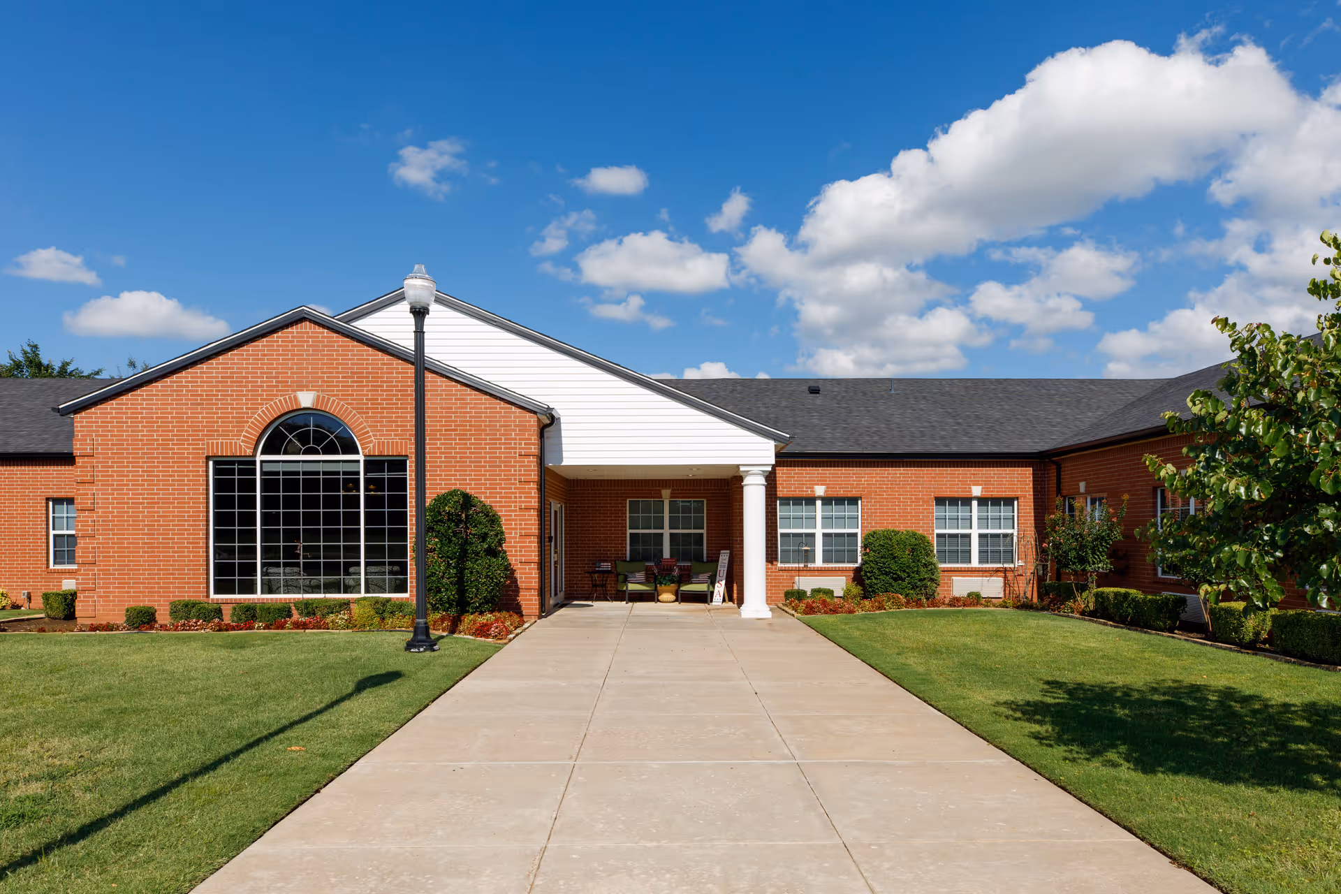 Front exterior view of a single-story brick building with a large arched window, white columns at the entrance, a concrete walkway leading up to the door, green lawn, and a blue sky with scattered clouds.