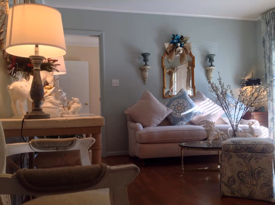 A cozy living room with a light pink sofa adorned with decorative pillows, a glass coffee table, and a patterned ottoman. The walls are painted light blue and decorated with a mirror and wall sconces. A table lamp and decorative figurines are placed on a side table, and natural light filters through a window with patterned curtains.