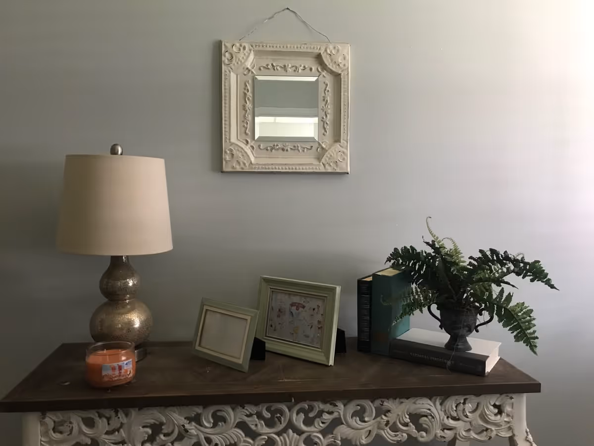 Decorative console table with a lamp, framed mirror, picture frames, books and a potted fern against a pale wall.