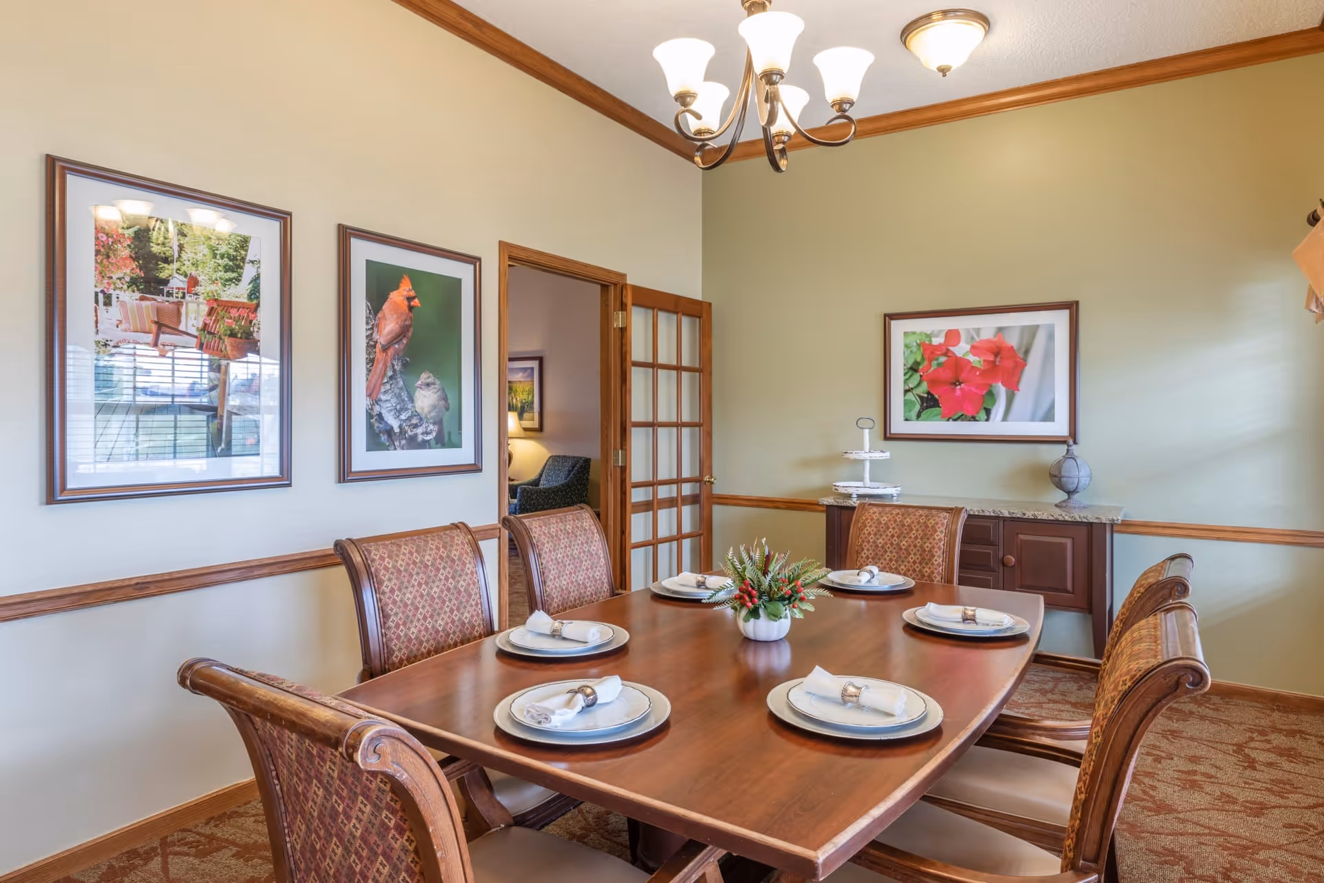 A dining room with a wooden table set for six people with plates, napkins, and napkin rings. The room has beige walls with wooden trim, three framed pictures on the walls, a chandelier overhead, and a sideboard with a decorative globe and tiered tray.
