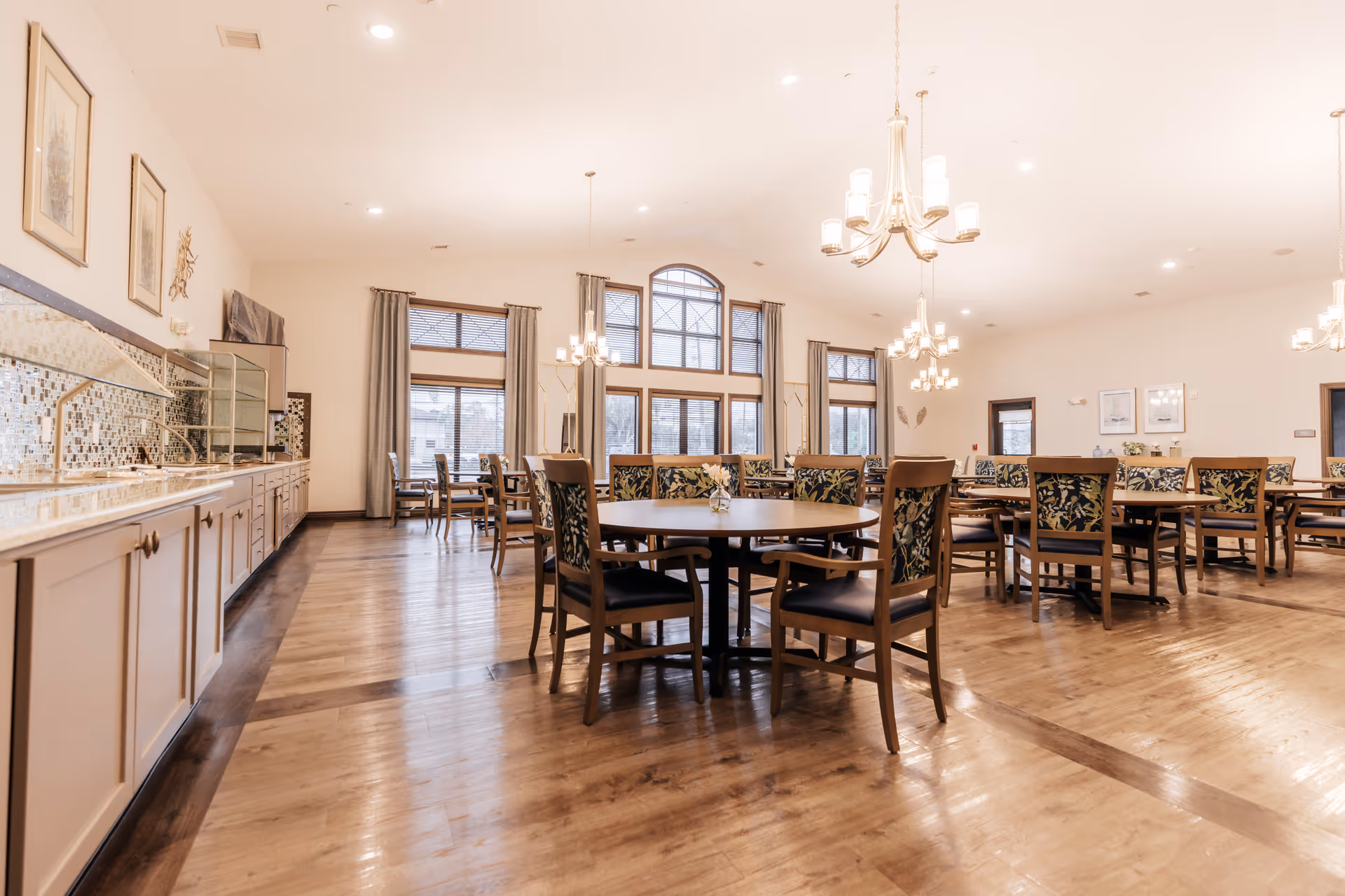 A spacious and well-lit dining room with multiple round and rectangular wooden tables surrounded by chairs with floral upholstery. The room features large windows with curtains, chandeliers hanging from the ceiling, and a long counter with cabinets and a mosaic tile backsplash along one wall.