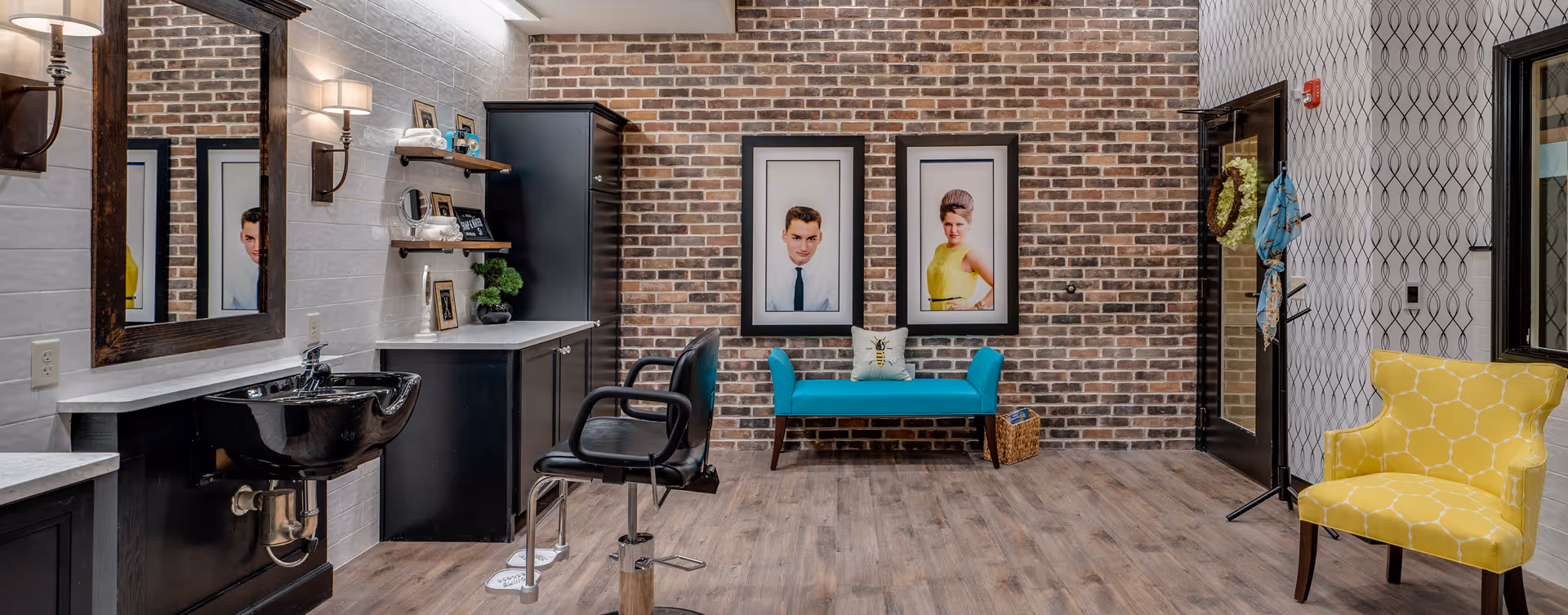 A bright interior salon area with a brick accent wall, two framed portraits above a turquoise bench, a yellow armchair, and a hair-washing/styling station.