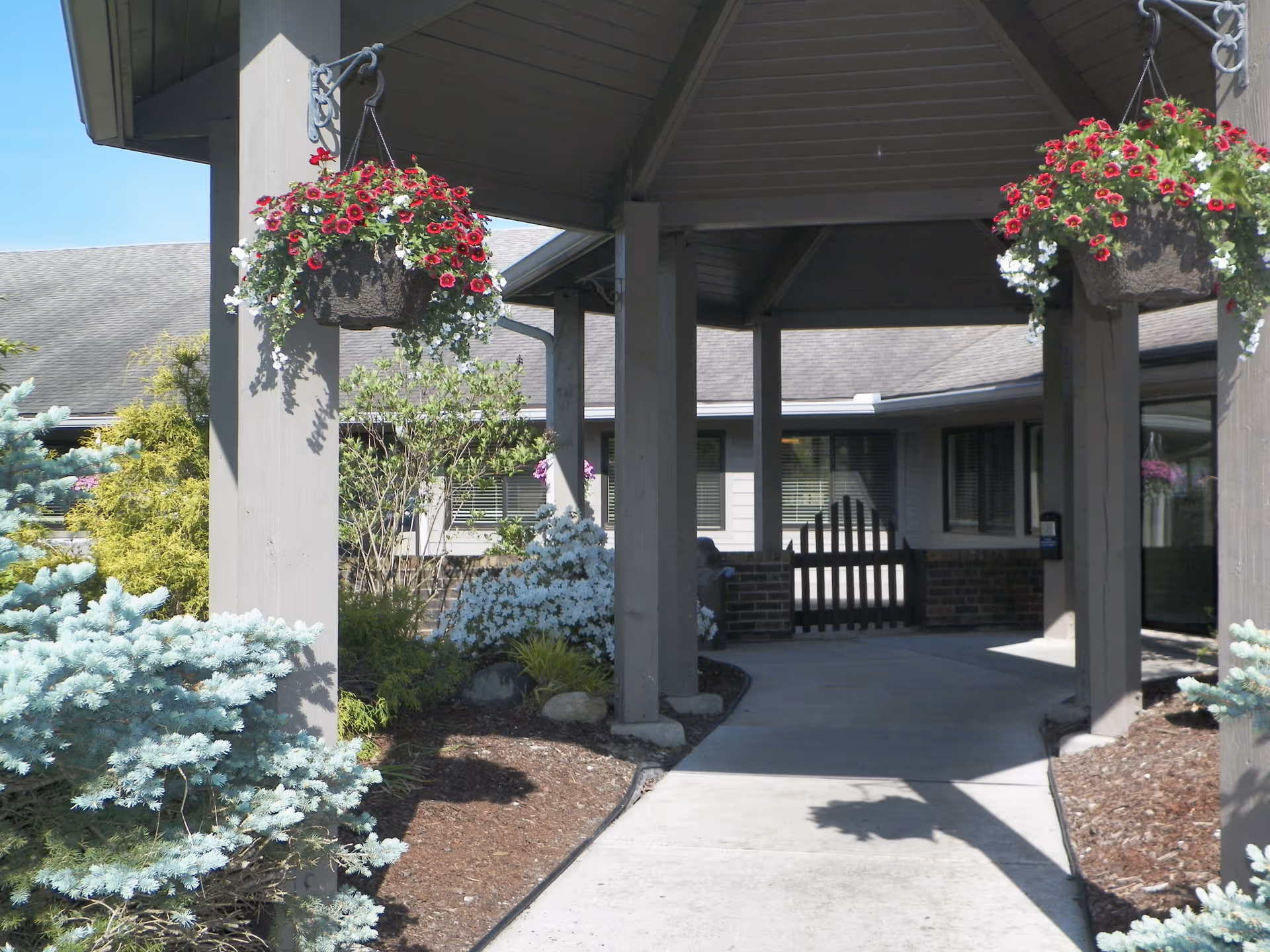 Covered walkway entrance to a building with hanging baskets of red and white flowers, surrounded by landscaped bushes and plants under a clear blue sky.