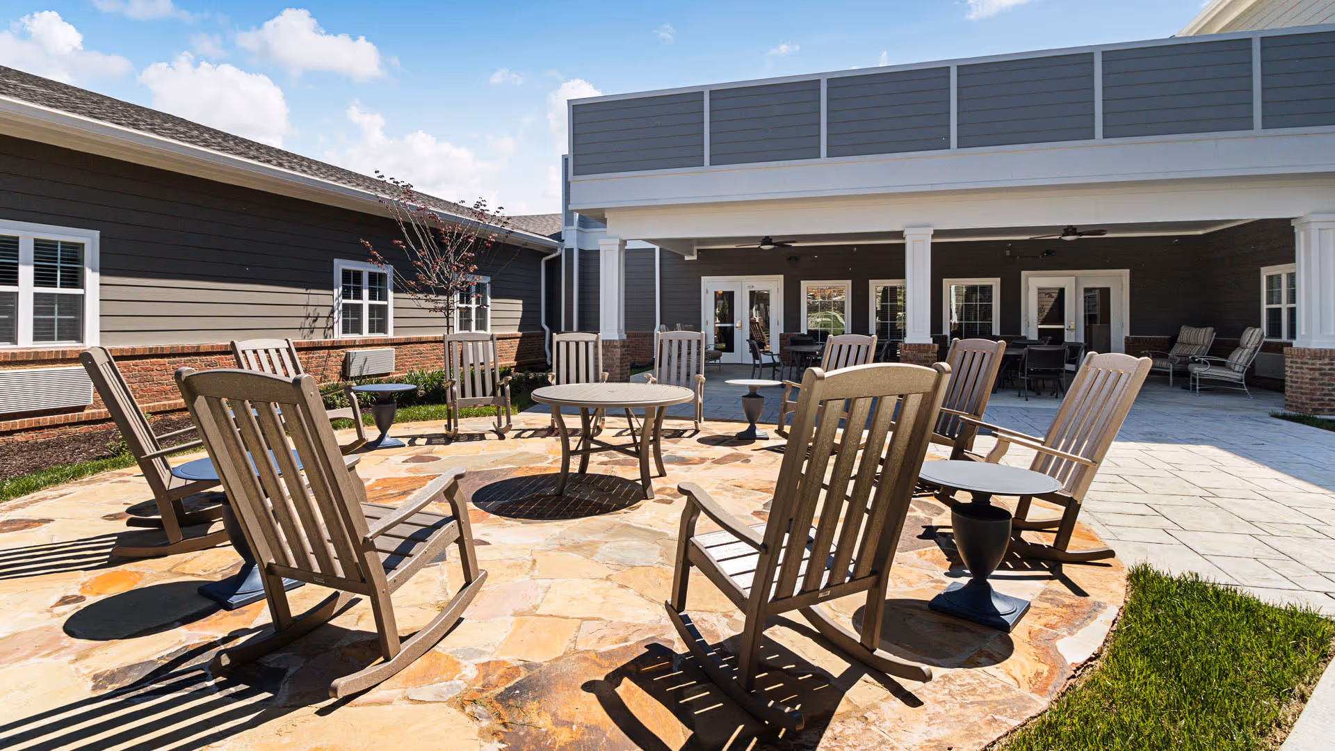Wooden rocking chairs and small tables arranged on a stone patio in front of a covered community building.