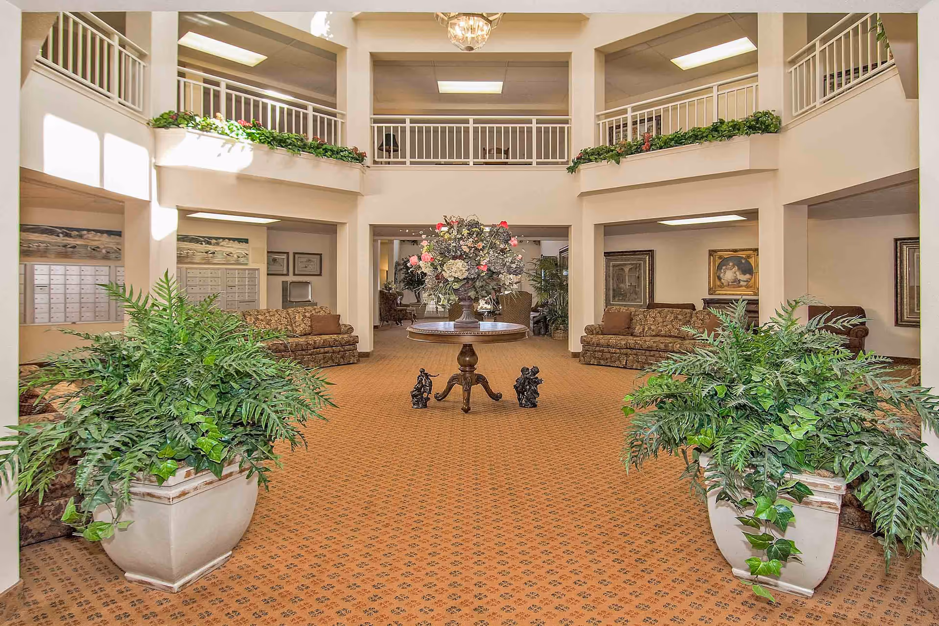 Bright two-story lobby with a central table topped by a floral arrangement, seating areas and large potted plants.