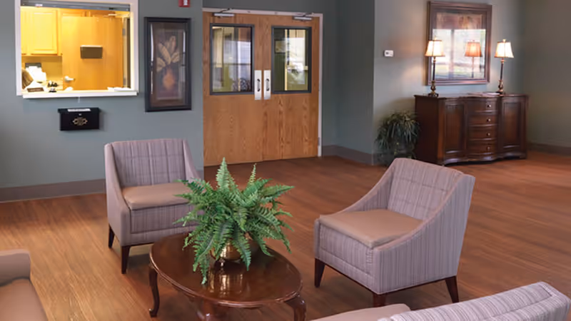 A cozy nursing home common area with wooden flooring, two beige armchairs, a wooden coffee table with a green potted fern, a wooden sideboard with three lamps and a framed picture above it, and double wooden doors with glass panels. There is also a reception window to the left with a view into a kitchen or office area.