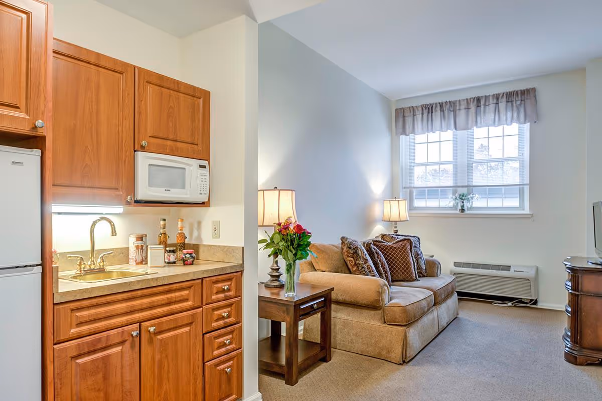 Small studio interior showing a kitchenette with wooden cabinets and a sink opening onto a cozy living area with a sofa, side table, lamps, and a window.