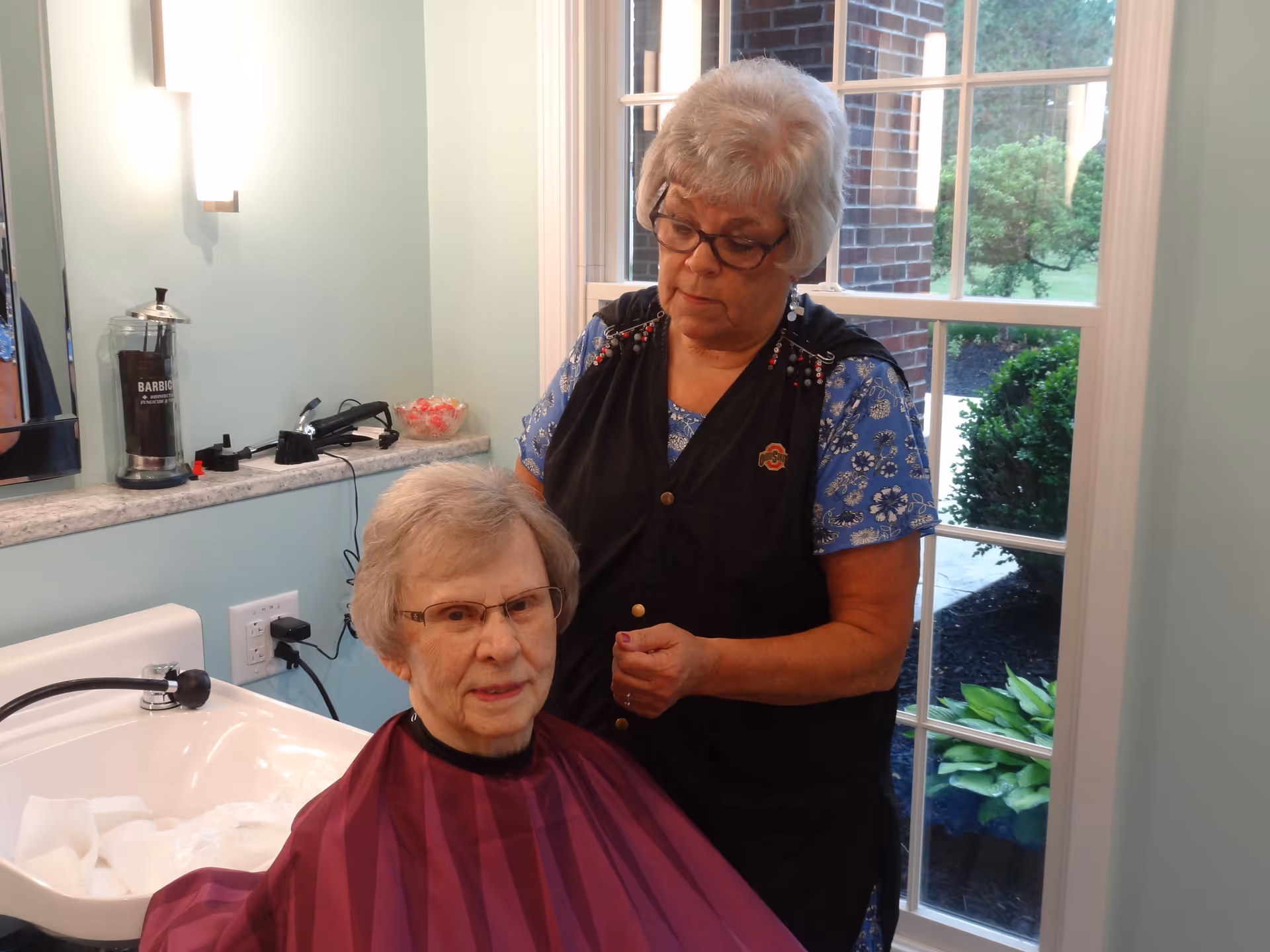 An elderly woman with glasses is seated wearing a maroon haircut cape while another woman stands behind her, appearing to style or cut her hair in a well-lit room with a large window showing greenery outside.