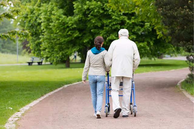 An elderly man using a walker is being assisted by a younger woman as they walk together on a paved path in a green park with trees and grass.