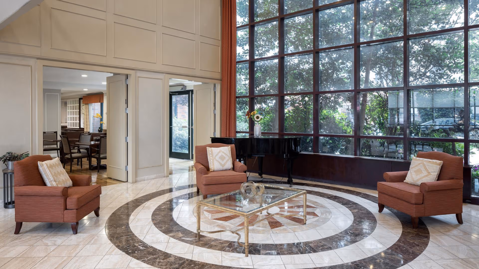 Sunlit lobby seating area with three upholstered chairs around a glass coffee table on a circular marble floor in front of a large window and a grand piano.