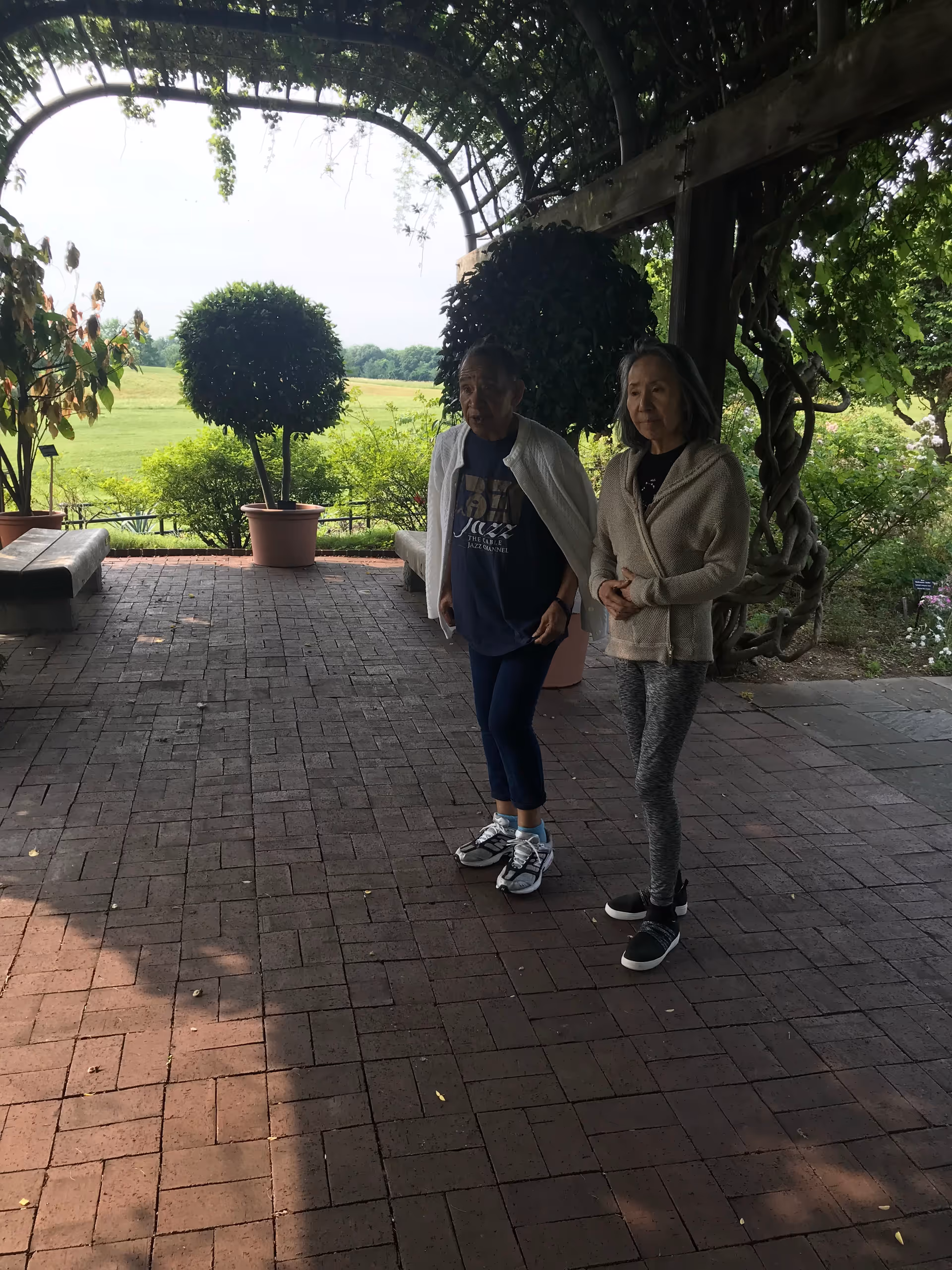 Two elderly women standing under a wooden pergola with climbing plants, overlooking a green outdoor garden area with potted plants and trees in the background.