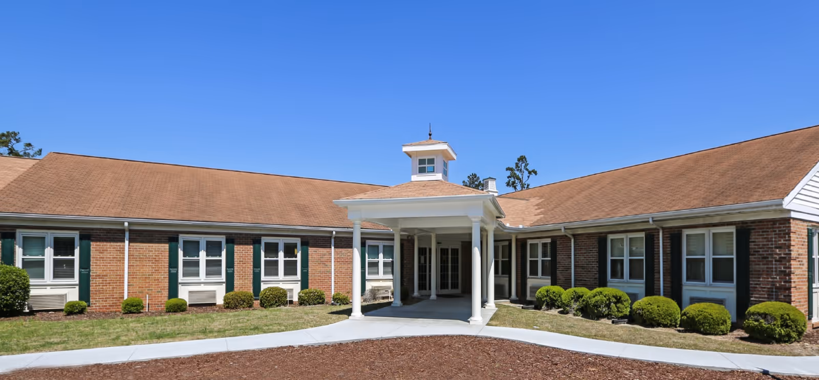 Exterior front view of a single-story brick building with a brown shingled roof and white columns at the entrance, surrounded by small bushes and a clear blue sky.