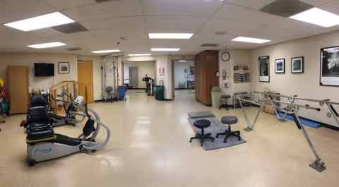 Wide view of a bright rehabilitation room with exercise machines, parallel bars, stools, and therapy equipment.