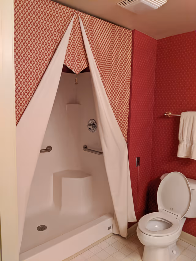 A bathroom with a white toilet and an open lid, a towel hanging on a silver towel rack, and a shower area with a built-in seat and grab bars. The shower is partially covered by a white curtain with a red and white patterned valance. The walls are covered in red wallpaper with a subtle pattern.