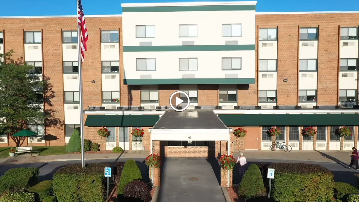 Front exterior of a multi-story brick senior living facility with a covered entrance, flagpole, hanging flower baskets and landscaped shrubs.