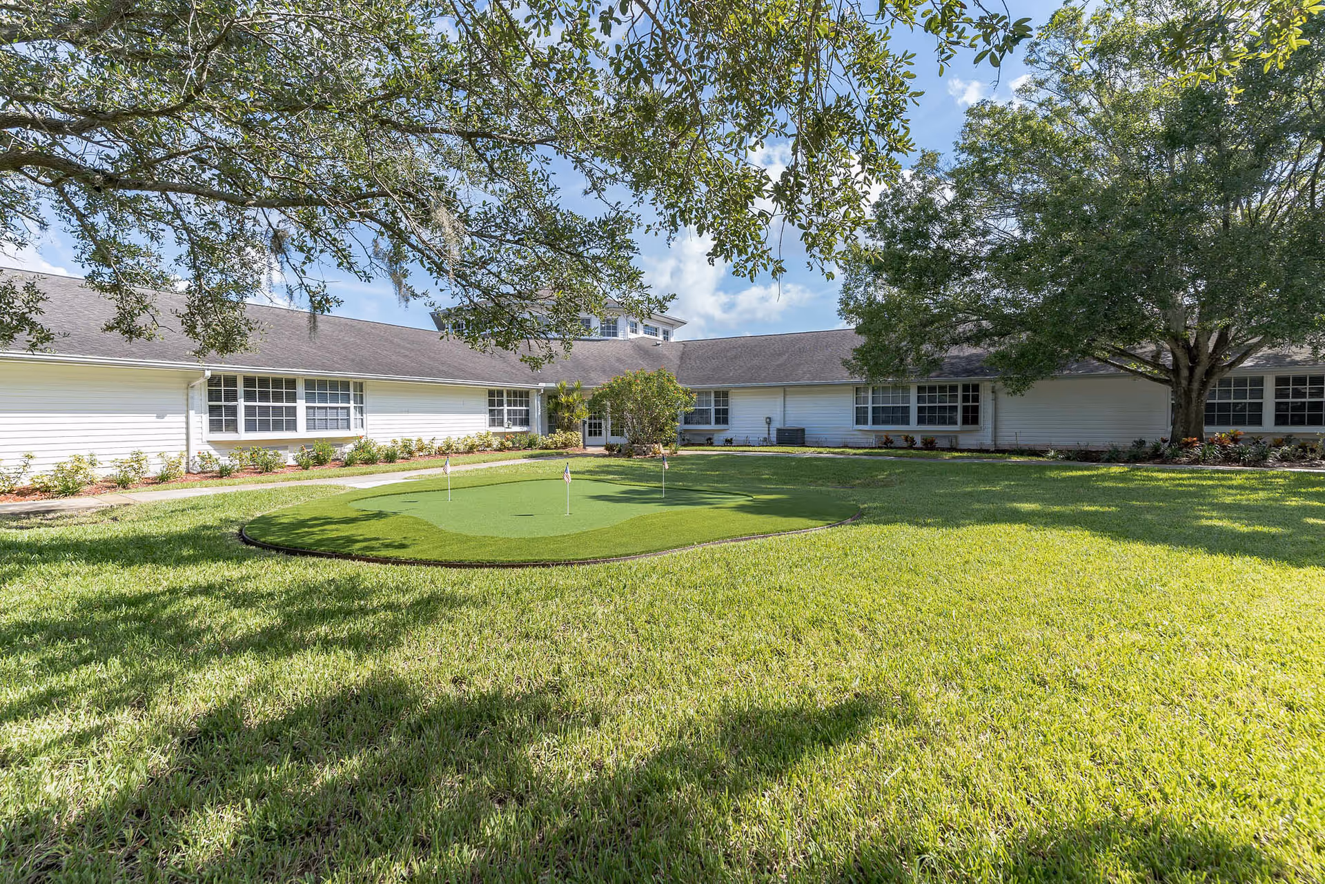 A sunny outdoor courtyard area with a small putting green surrounded by well-maintained grass and trees. The courtyard is enclosed by a single-story white building with multiple windows and a shingled roof. Branches of trees frame the top of the image, casting shadows on the grass.