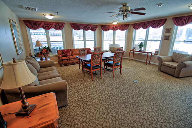 A spacious living room with large windows covered by red valances, featuring a brown sofa, two armchairs, a wooden table with four chairs, a side table with a lamp, and decorative plants and framed pictures on the walls.