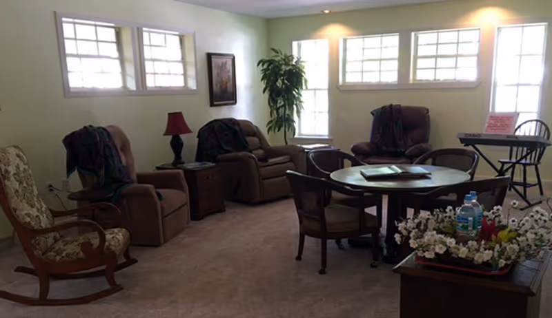A cozy living room with three recliner chairs, a floral rocking chair, a round wooden table with four chairs, a side table with a red lamp, a potted plant near the window, a keyboard on a stand, and a small table with a floral arrangement and a water bottle.