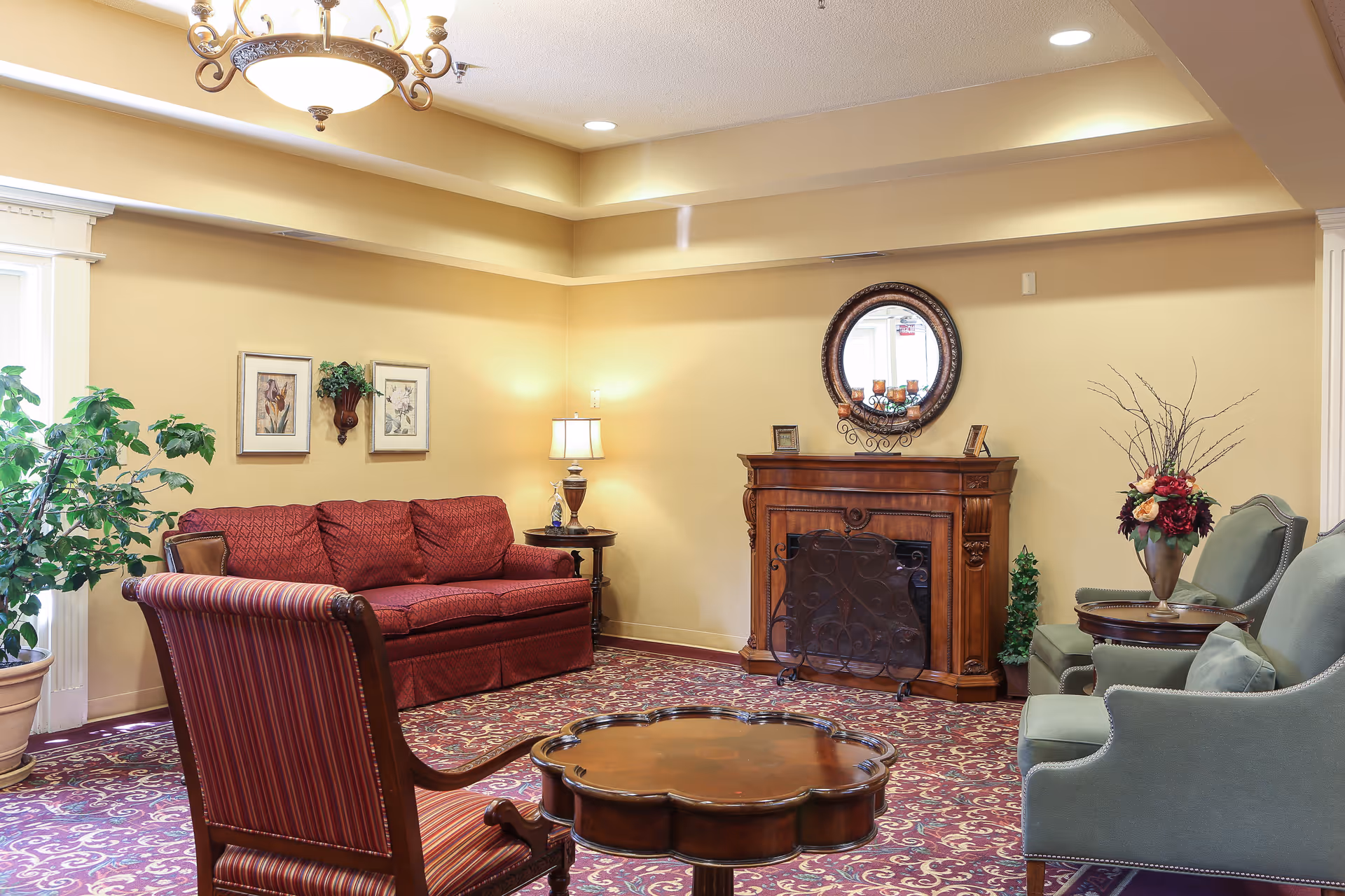 A cozy living room area with a red patterned sofa, two green armchairs, a wooden coffee table, and a wooden fireplace with a round mirror above it. The room has beige walls, a patterned carpet, a potted plant, framed artwork, and a decorative lamp on a side table.
