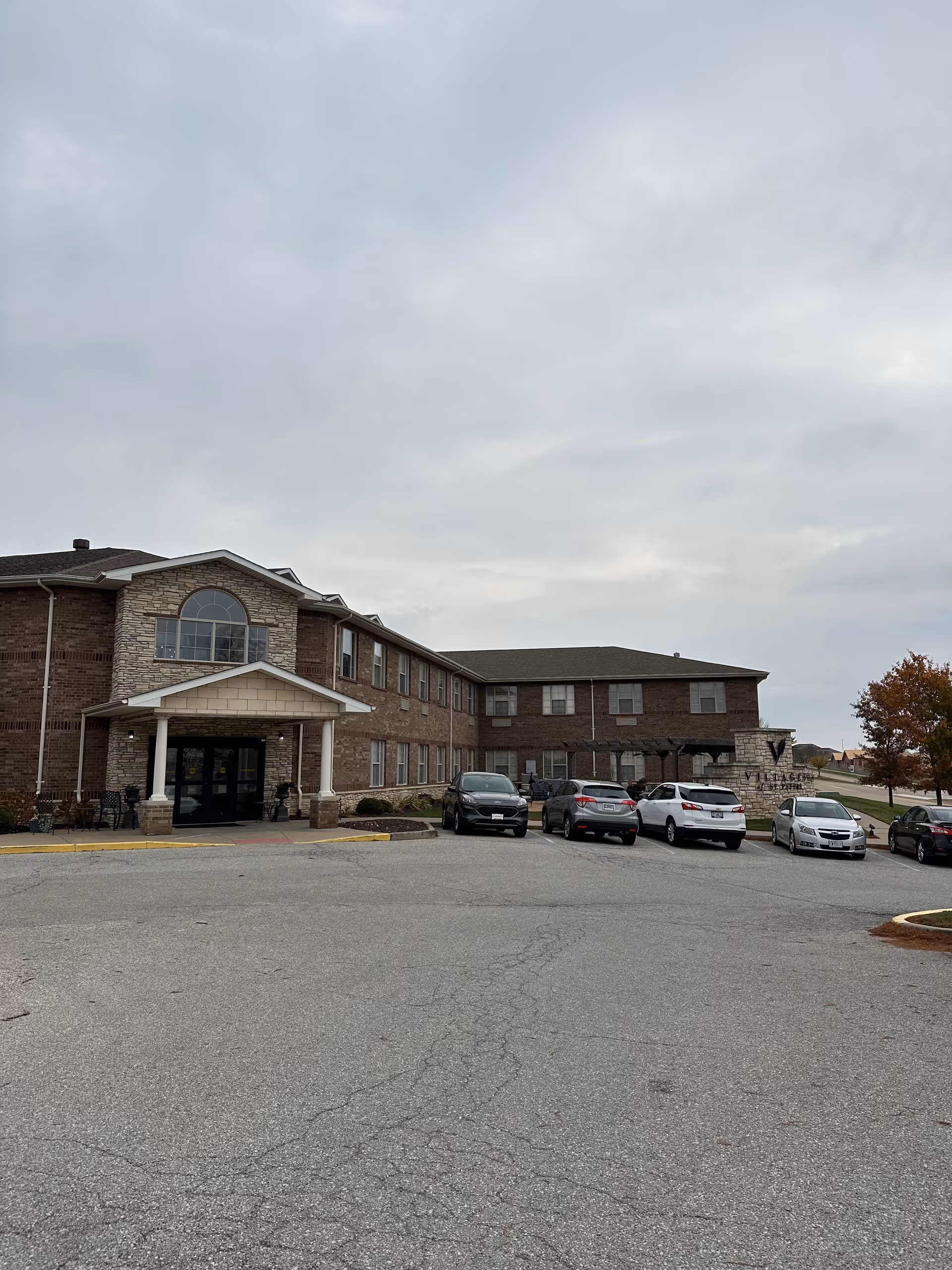 Two-story brick senior living building with a covered entrance and several cars parked in the front lot under an overcast sky.