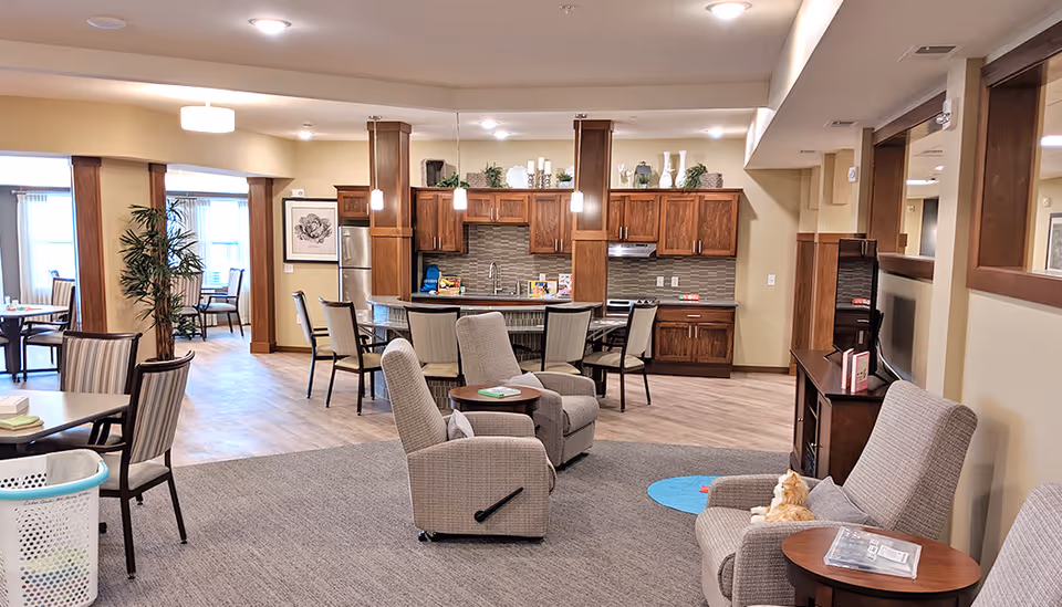 A spacious senior living common area featuring a kitchen with wooden cabinets, a refrigerator, and a stove. In front of the kitchen is a dining area with tables and chairs. The foreground has comfortable armchairs and small tables, one with a cat sitting on it. The room is well-lit with ceiling lights and pendant lights over the kitchen island. There are plants and decorative items on top of the cabinets and framed artwork on the walls.
