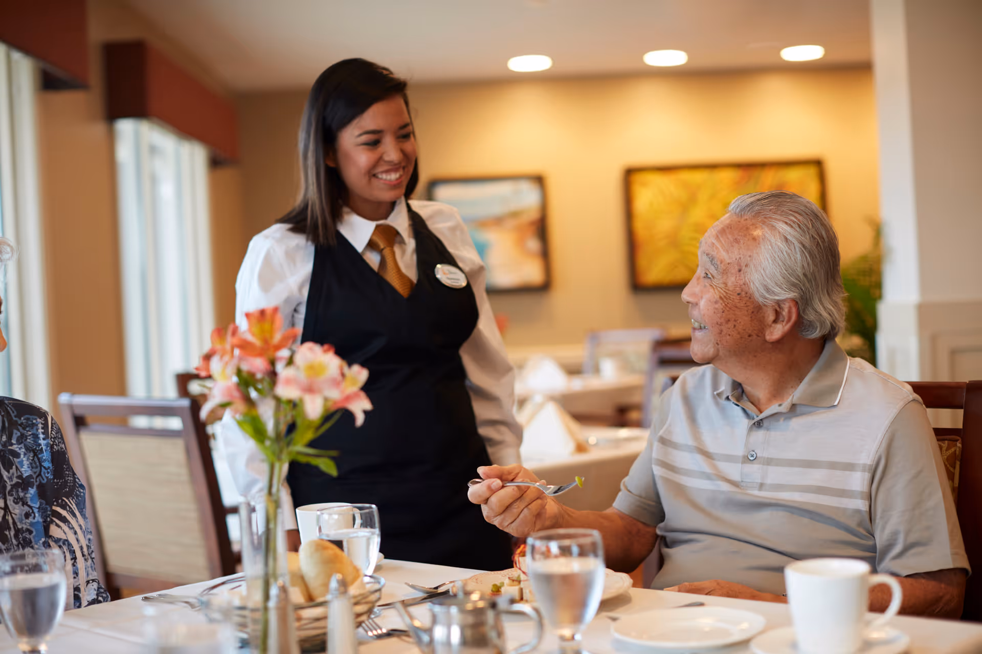 A smiling waitress in a black vest and white shirt interacts with an elderly man seated at a dining table in a well-lit dining room. The table is set with glasses of water, a bread roll, and a flower vase with pink flowers. The background shows framed artwork on the wall.