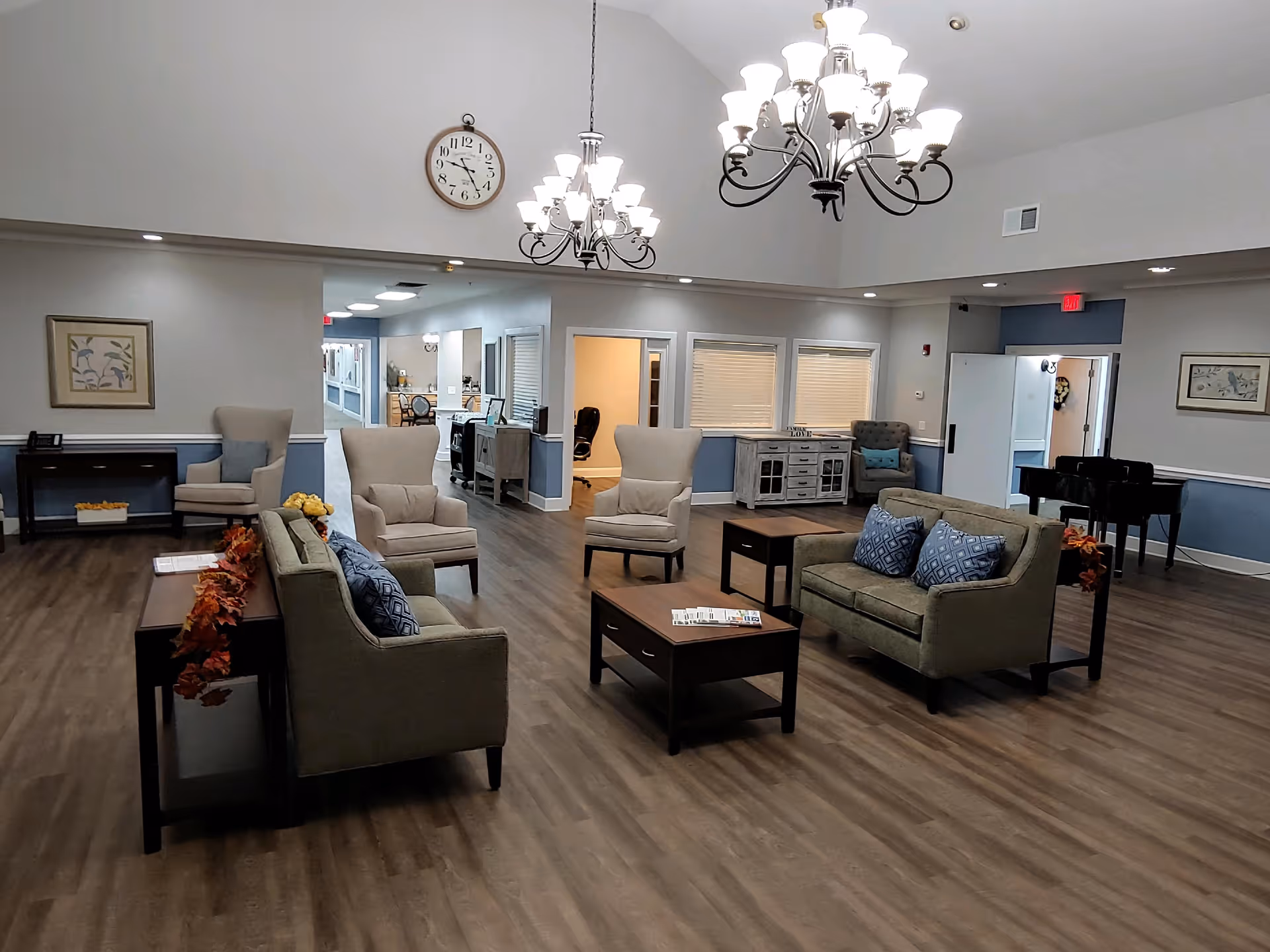 A spacious and well-lit living room area in a senior living facility with multiple armchairs and sofas arranged around two wooden coffee tables. The room features wood flooring, two large chandeliers, a wall clock, framed artwork, and a piano in the corner. The walls are painted in light gray and blue tones with white trim.