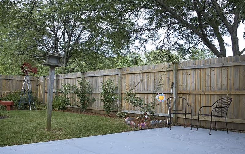 Fenced backyard patio with a lawn, birdhouse on a post, two metal chairs, and garden decorations.