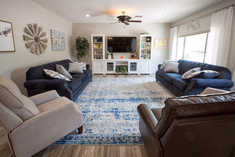 Bright living room with two navy sofas, two recliners, a large blue patterned rug, and a TV console with a wall-mounted television.