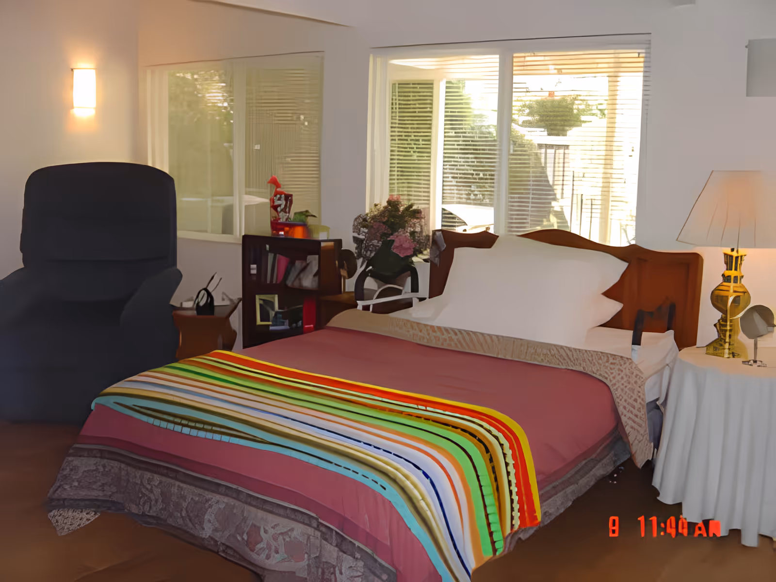 A furnished bedroom with a made bed covered by a colorful striped blanket, a recliner, bedside tables with lamps, and a window with blinds.