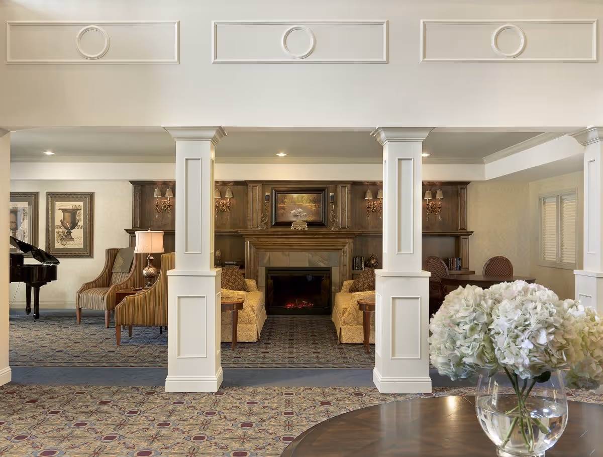 A cozy living room area in a senior living facility featuring two white columns framing a view of a fireplace with a wooden mantel and a painting above it. There are two beige armchairs with patterned cushions on either side of the fireplace, a pair of striped armchairs with a lamp between them to the left, and a grand piano in the far left corner. A round table with a glass vase of white hydrangeas is in the foreground on a patterned carpeted floor.