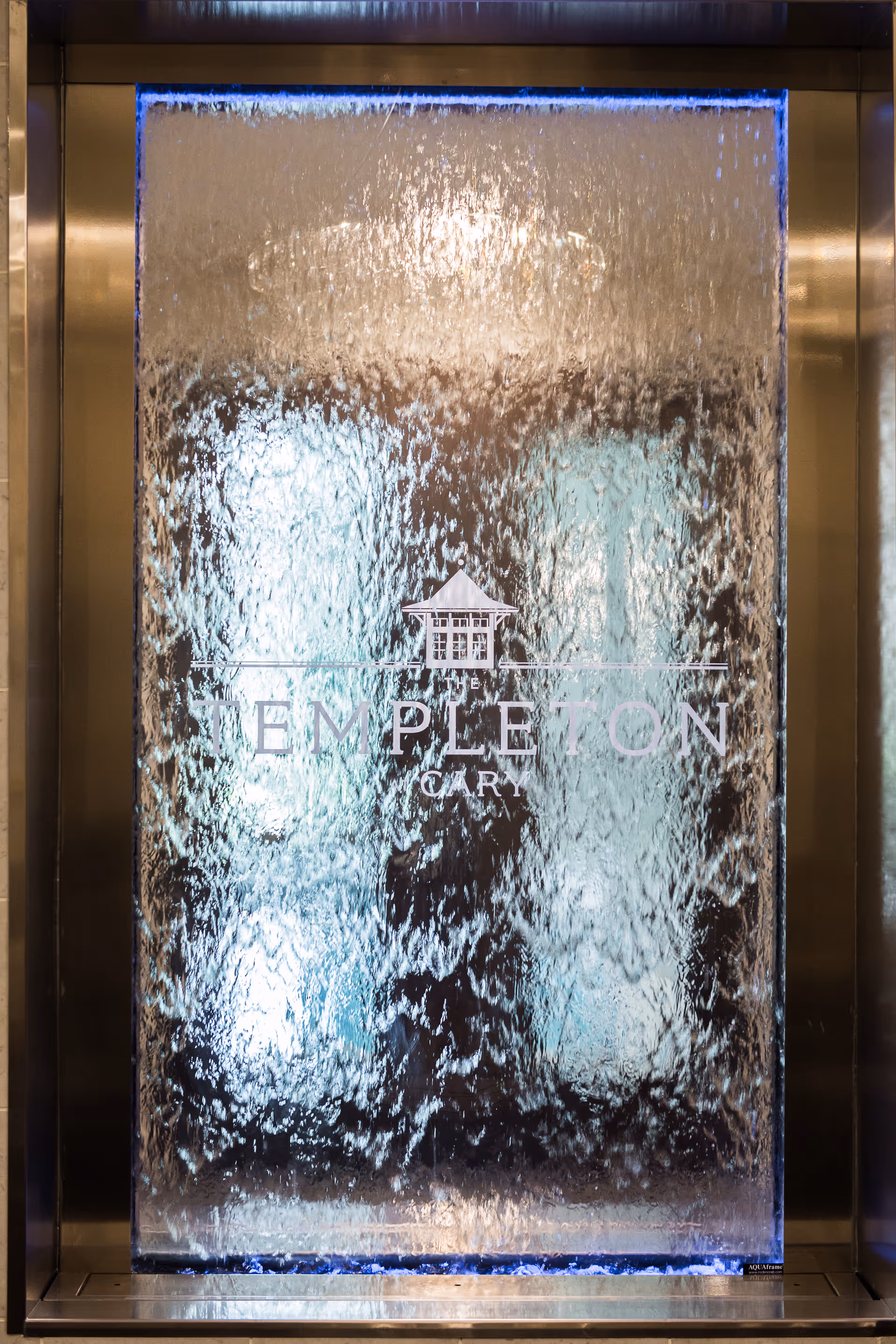 A decorative indoor water wall feature with water cascading down a glass panel. The glass panel has the logo and text 'The Templeton Cary' etched on it, framed by a metallic border with blue lighting at the top and bottom edges.