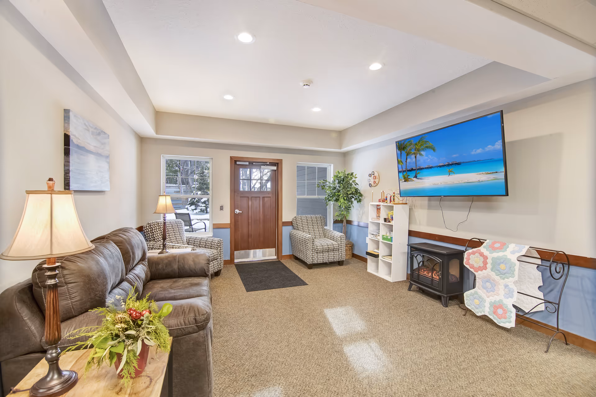 A cozy living room area with a brown leather sofa, two patterned armchairs, a wooden door with windows on either side, a flat-screen TV mounted on the wall displaying a beach scene, a small electric fireplace, a white shelving unit with books and games, a quilt rack with a colorful quilt, a floor lamp, and a potted plant.