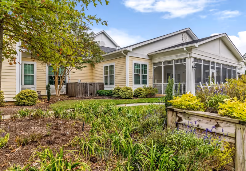 Exterior view of a single-story building with beige siding and white trim, surrounded by a garden with various green plants and flowers. There is a screened porch on the right side and a tree with green leaves on the left. The sky is partly cloudy.