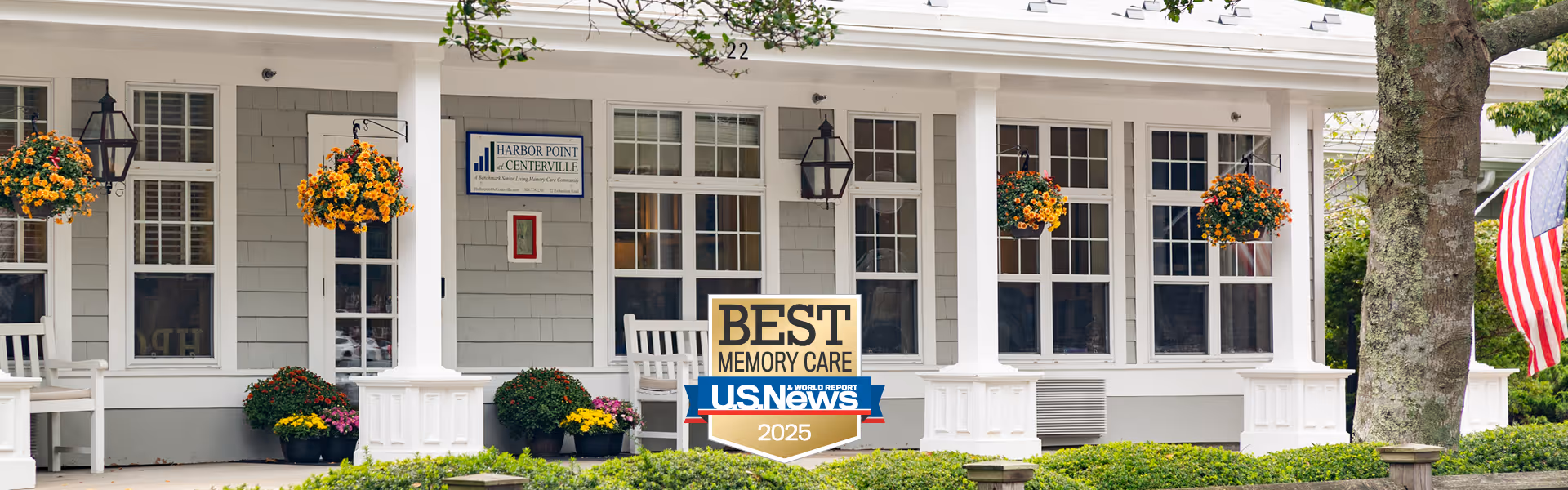 Front exterior view of Harbor Point at Centerville facility showing a porch with white rocking chairs, hanging flower baskets with orange and yellow flowers, potted plants, and an American flag on the right side. The building has gray siding, white trim, and multiple windows. A sign on the building reads 'Harbor Point at Centerville'.