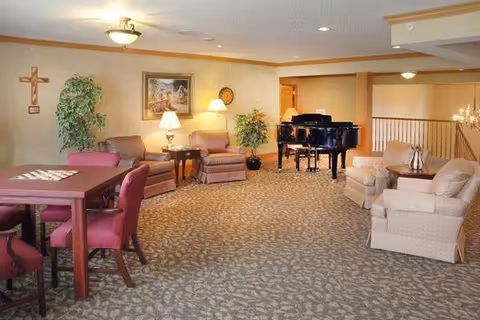 A spacious living room area in a senior living facility with patterned carpet, a wooden table with four red upholstered chairs, two armchairs with a side table and lamp, a sofa with a side table and lamp, potted plants, a wall cross, a framed painting, and a black grand piano in the background.