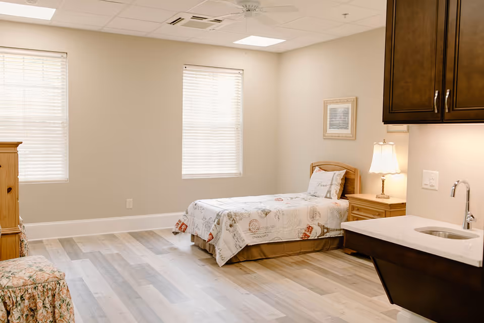 A bright and clean assisted living bedroom with a single bed covered in a patterned quilt, a wooden nightstand with a lamp, two windows with blinds, a wooden dresser, a floral upholstered chair, and a small sink with a countertop and dark wood cabinets.
