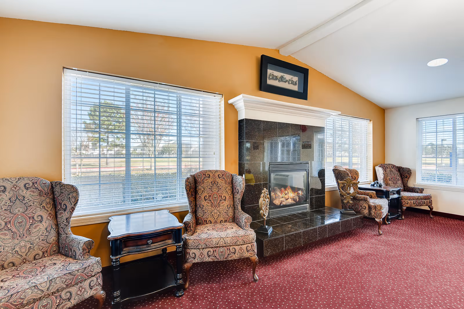 A cozy living room area with four patterned armchairs arranged around a black tiled fireplace with a fire burning. There are two wooden side tables between the chairs, and large windows with white blinds letting in natural light. The walls are painted a warm yellow and white, and the floor is covered with a red carpet with a subtle pattern.