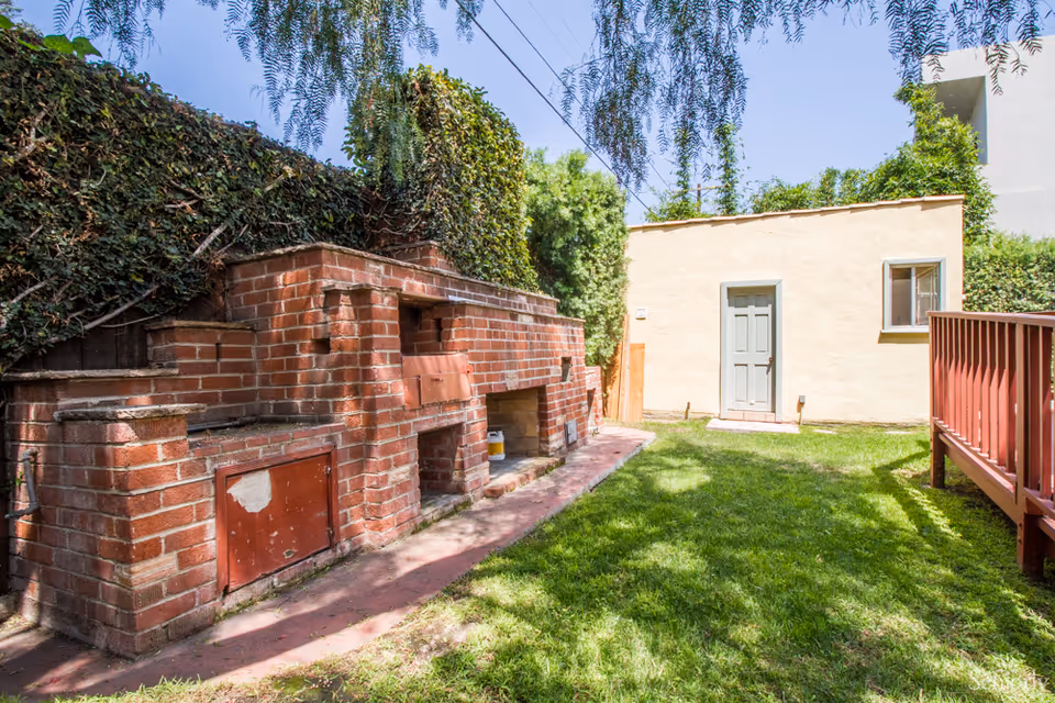 Outdoor area with a brick barbecue grill and oven setup along a wall covered with ivy. There is a grassy lawn leading to a small beige building with a gray door and window. A wooden deck with red railing is visible on the right side under a clear blue sky.