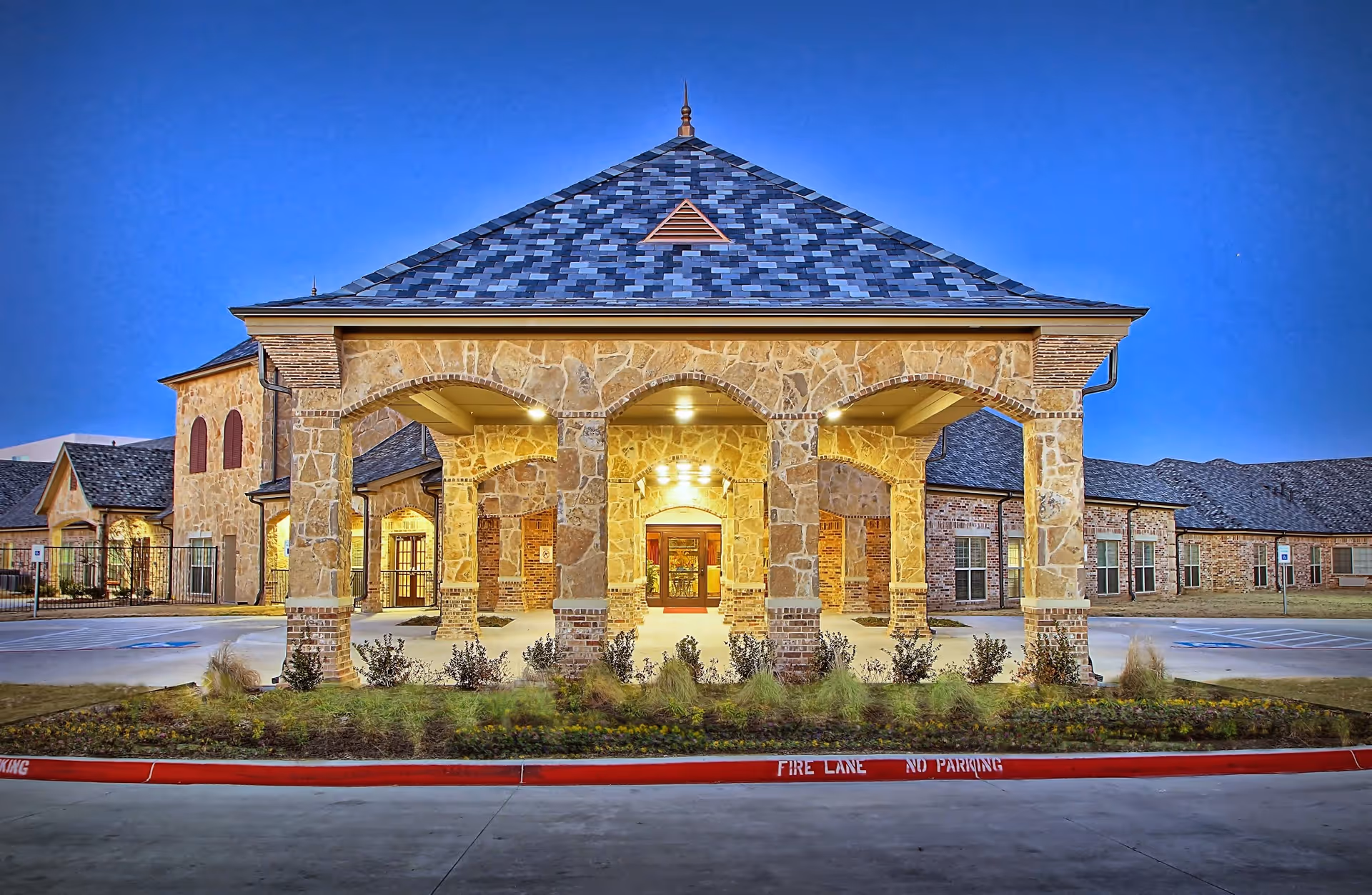 Front exterior view of Accel at Willow Bend facility at dusk, featuring a stone and brick entrance with arches and a peaked roof, surrounded by a parking area and landscaped greenery.