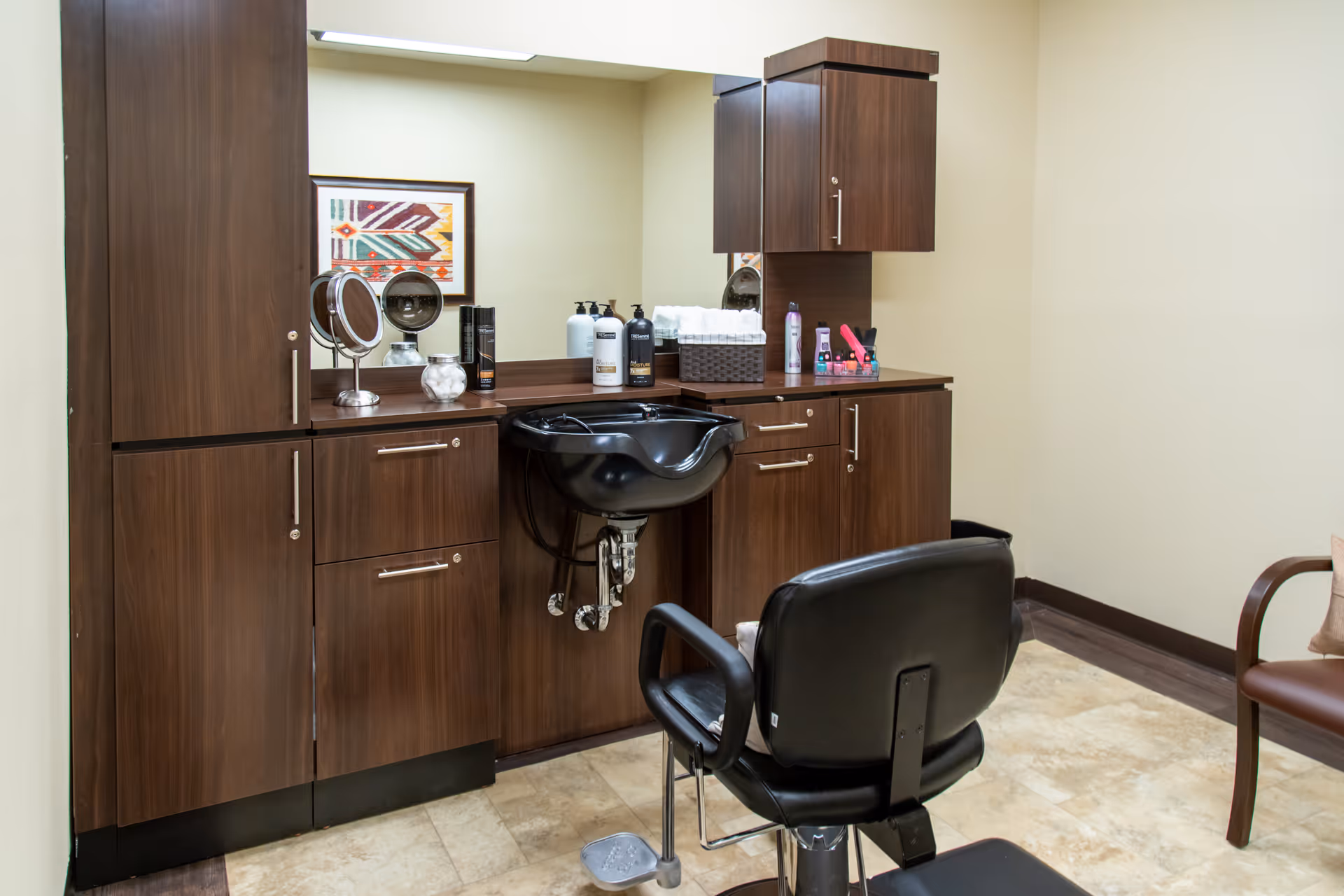 Interior salon station with a black styling chair, shampoo sink, and dark wood cabinets and mirror.