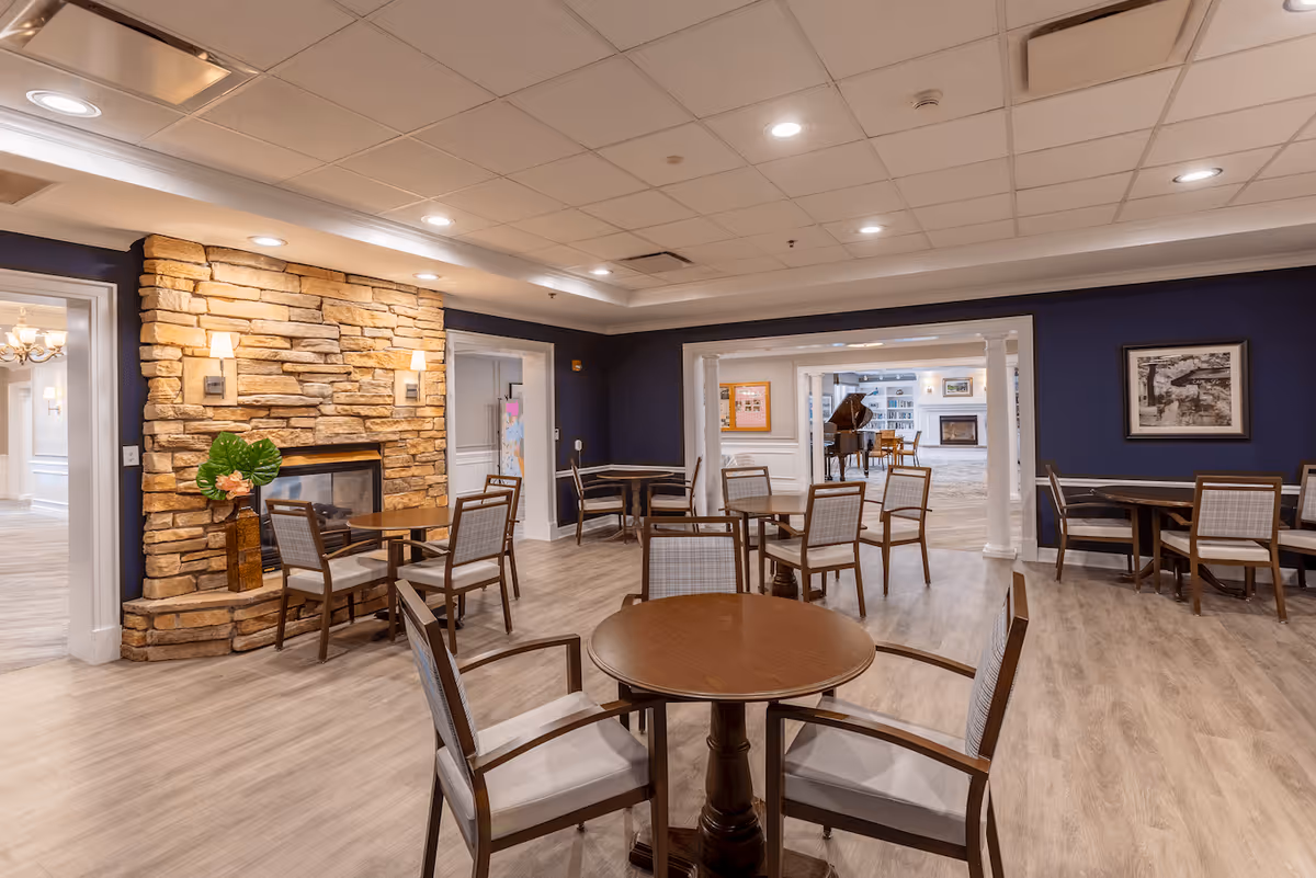 A cozy interior room with multiple round wooden tables and chairs arranged for seating. The room features a stone fireplace with two wall sconces and a decorative plant on the mantel. The walls are painted dark blue with white trim, and the floor has light wood-style flooring. Through an open doorway, another room with a grand piano and additional seating is visible.
