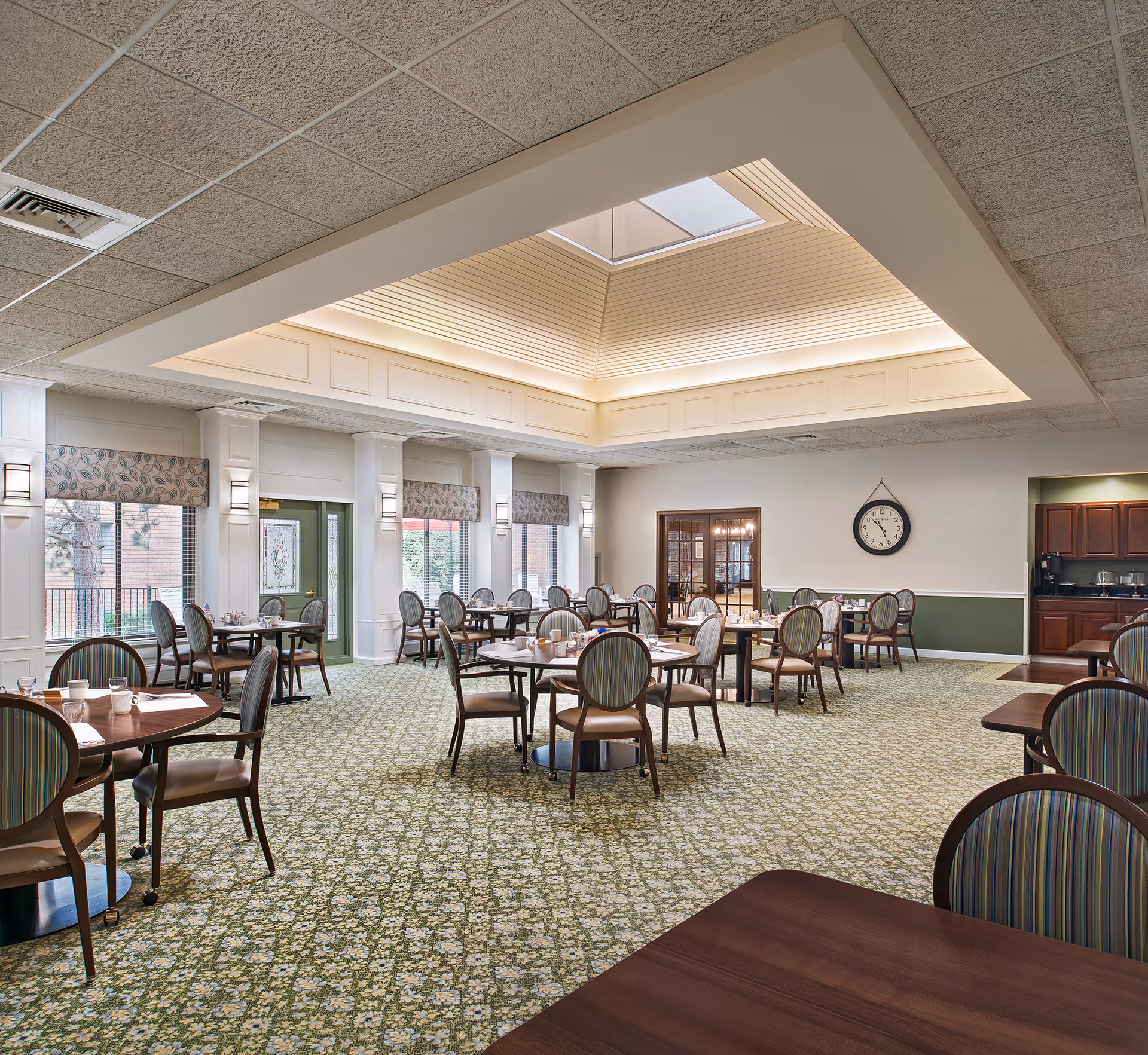 A spacious dining room with multiple round tables and chairs arranged neatly. The room features a patterned green carpet, large windows with floral valances, and a skylight in the ceiling providing natural light. A clock is mounted on the wall, and there is a small kitchenette area with wooden cabinets in the background.