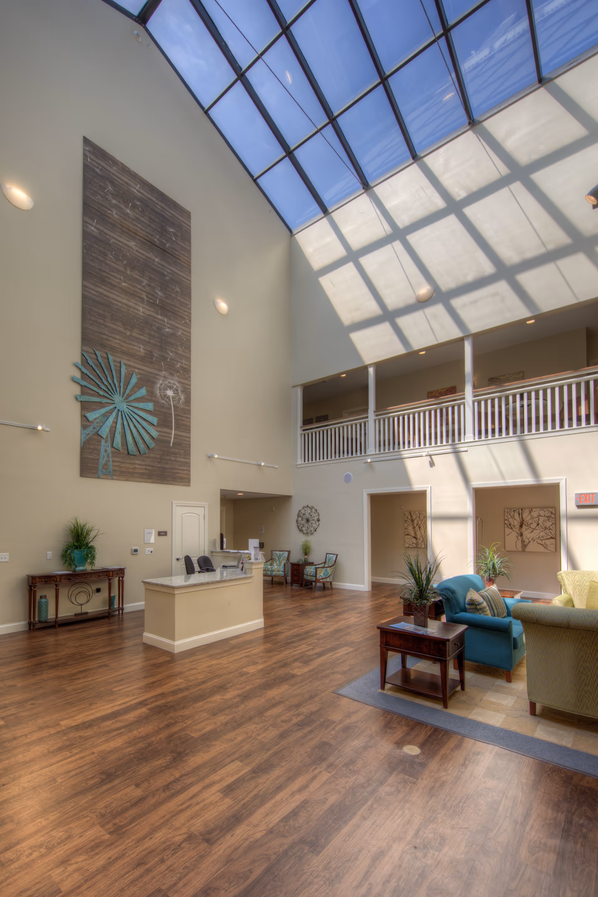 A spacious senior living facility common area with high ceilings and a large skylight letting in natural light. The room features wood flooring, a reception desk, seating area with armchairs and a small table, decorative plants, and wall art including a large piece with a windmill design.