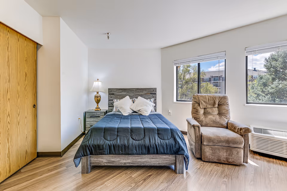 A bright senior living bedroom with a wooden bed frame, dark blue bedding, two white pillows, a wooden nightstand with a decorative lamp, a comfortable brown recliner chair, two large windows showing trees and a building outside, and a wooden closet door on the left.