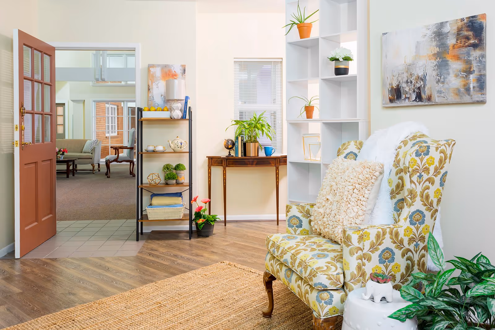 A cozy living room area with a floral patterned armchair adorned with a fluffy white throw and a textured beige pillow. Next to the chair is a small white side table with a small plant on it. Behind the chair is a white shelving unit with decorative plants and items. A wooden console table with plants and a globe sits against the wall under a window with blinds. To the left, an open door leads to another room with additional seating and a coffee table. The floor is a combination of wood and tile with a woven rug in the foreground. A colorful abstract painting hangs on the wall above the chair.
